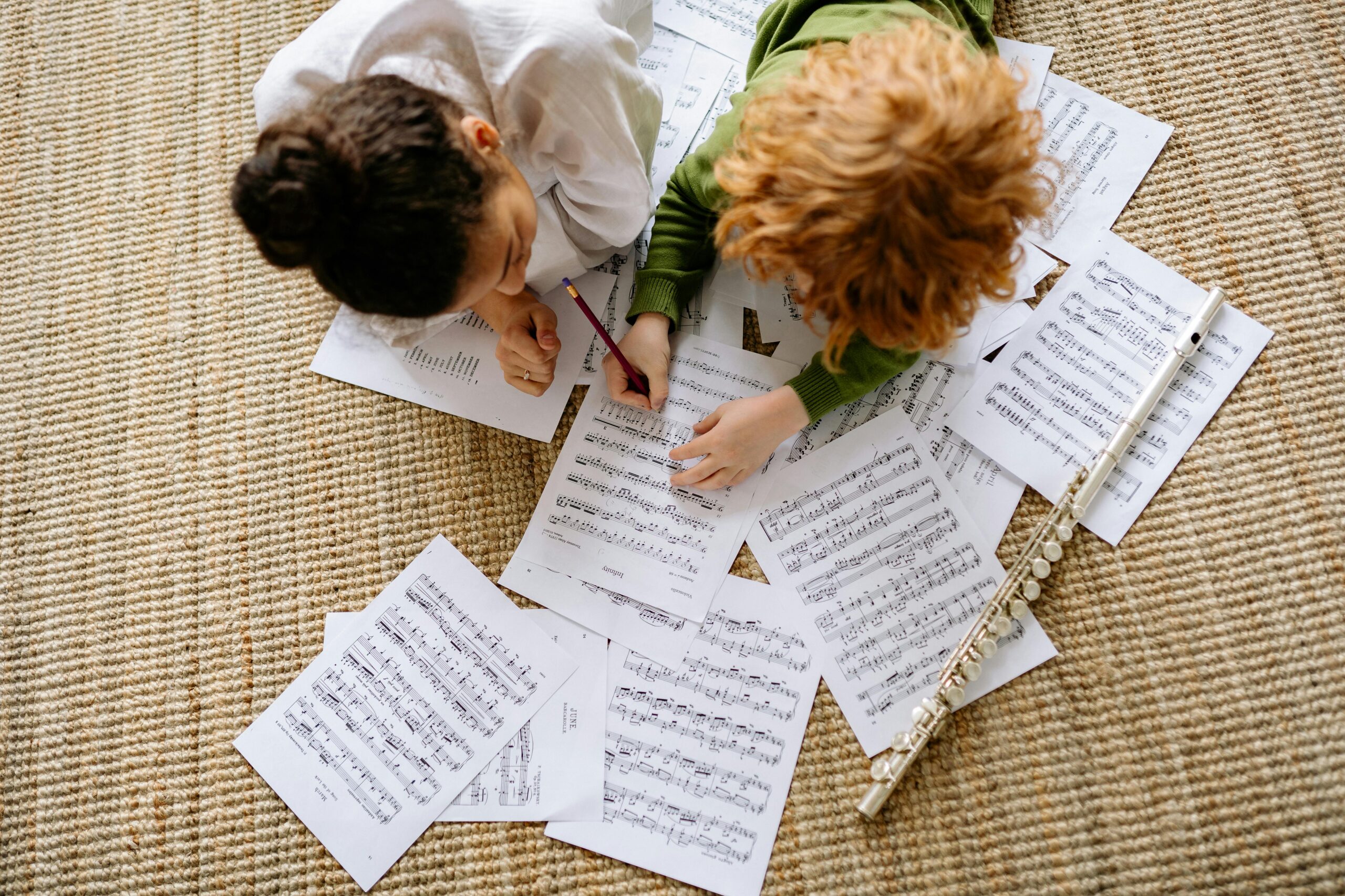 Two children learning music together with a flute and scattered sheet music on the floor.